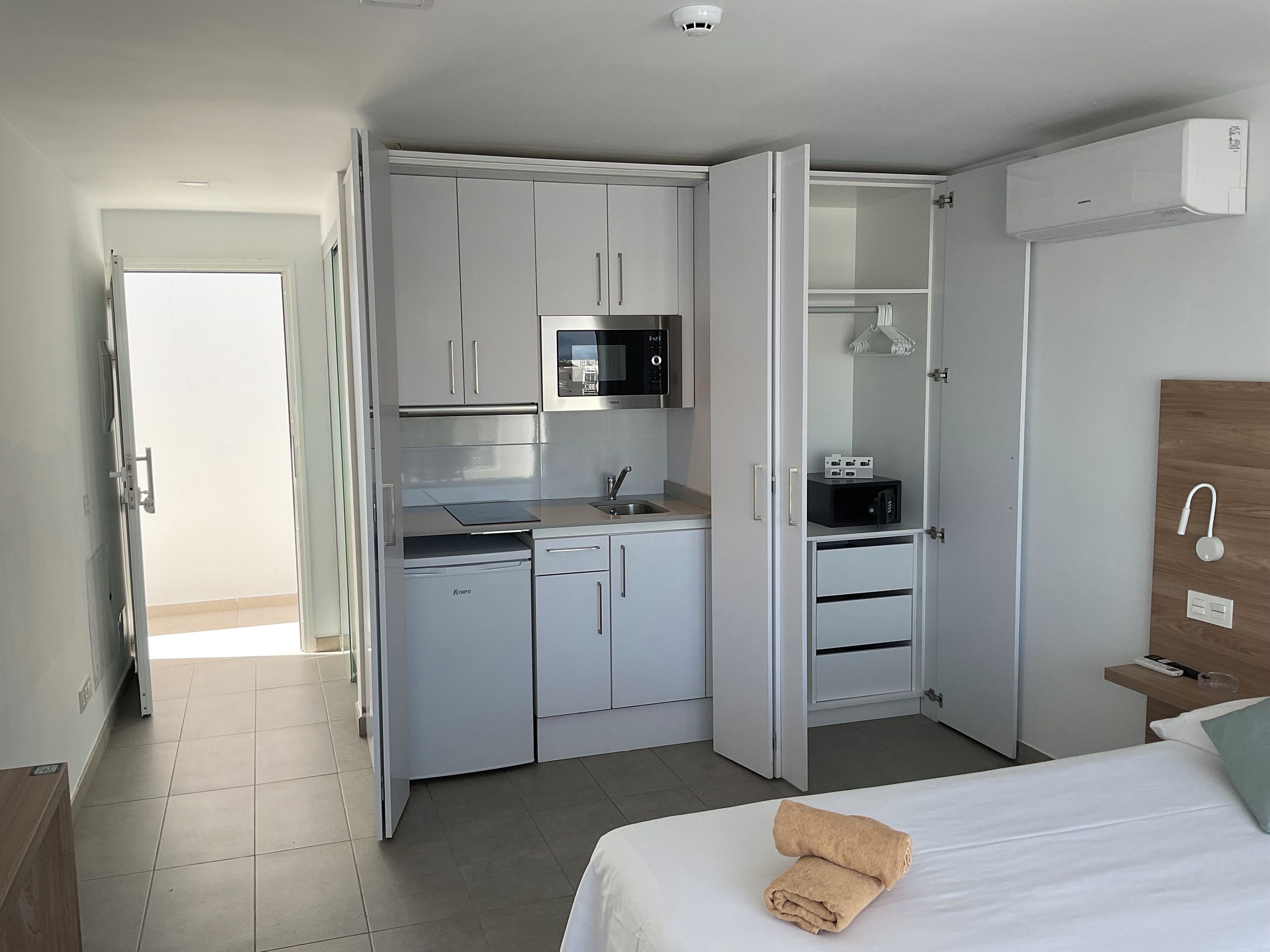 Ett kompakt hotellrum med ett litet pentry, en säng med en hoprullad handduk och en sänggavel i trä.