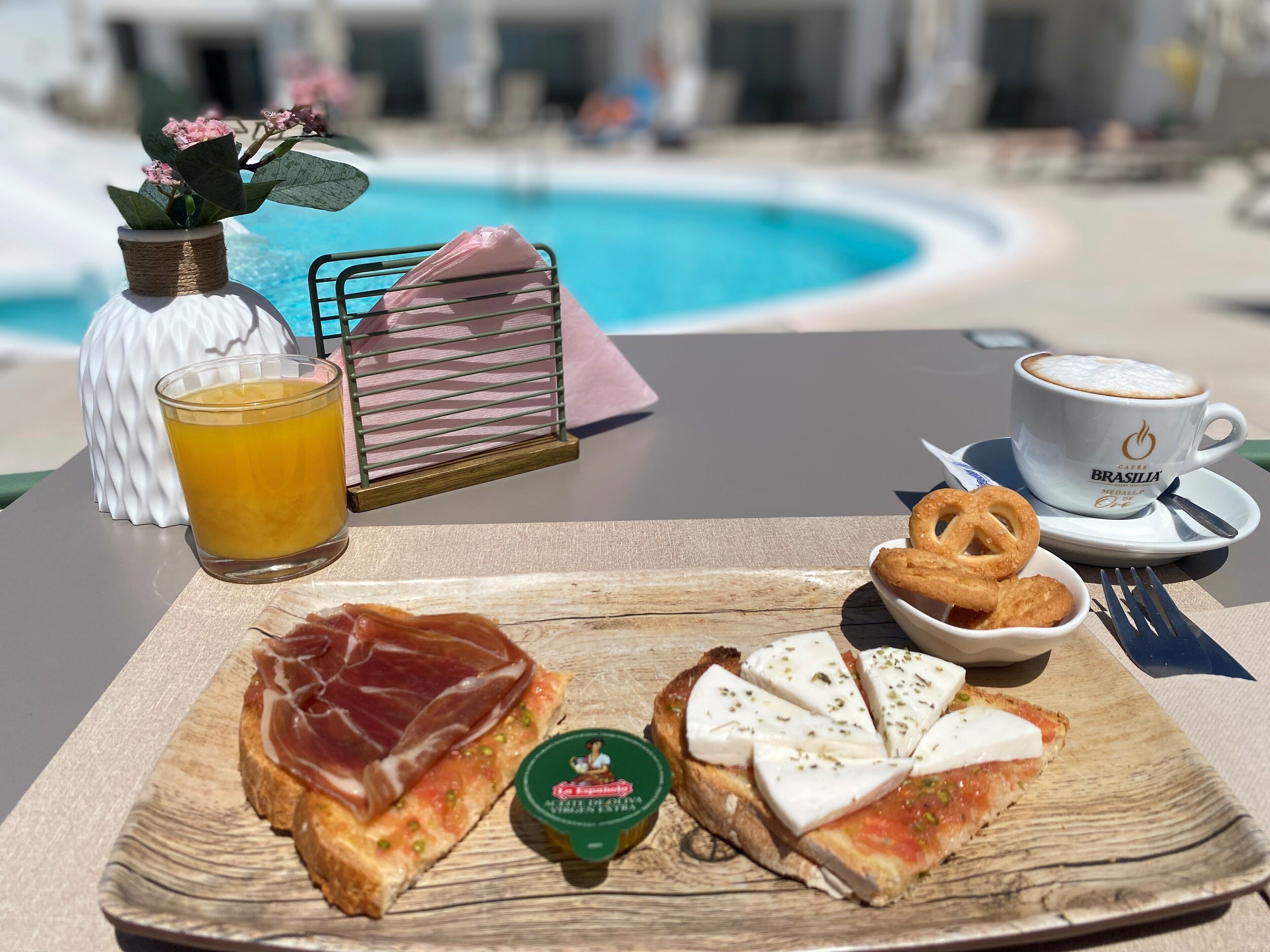 A breakfast tray with prosciutto, mozzarella, and a small jar of jam, a glass of orange juice, a cup of coffee, pretzels, and a napkin holder by a pool.