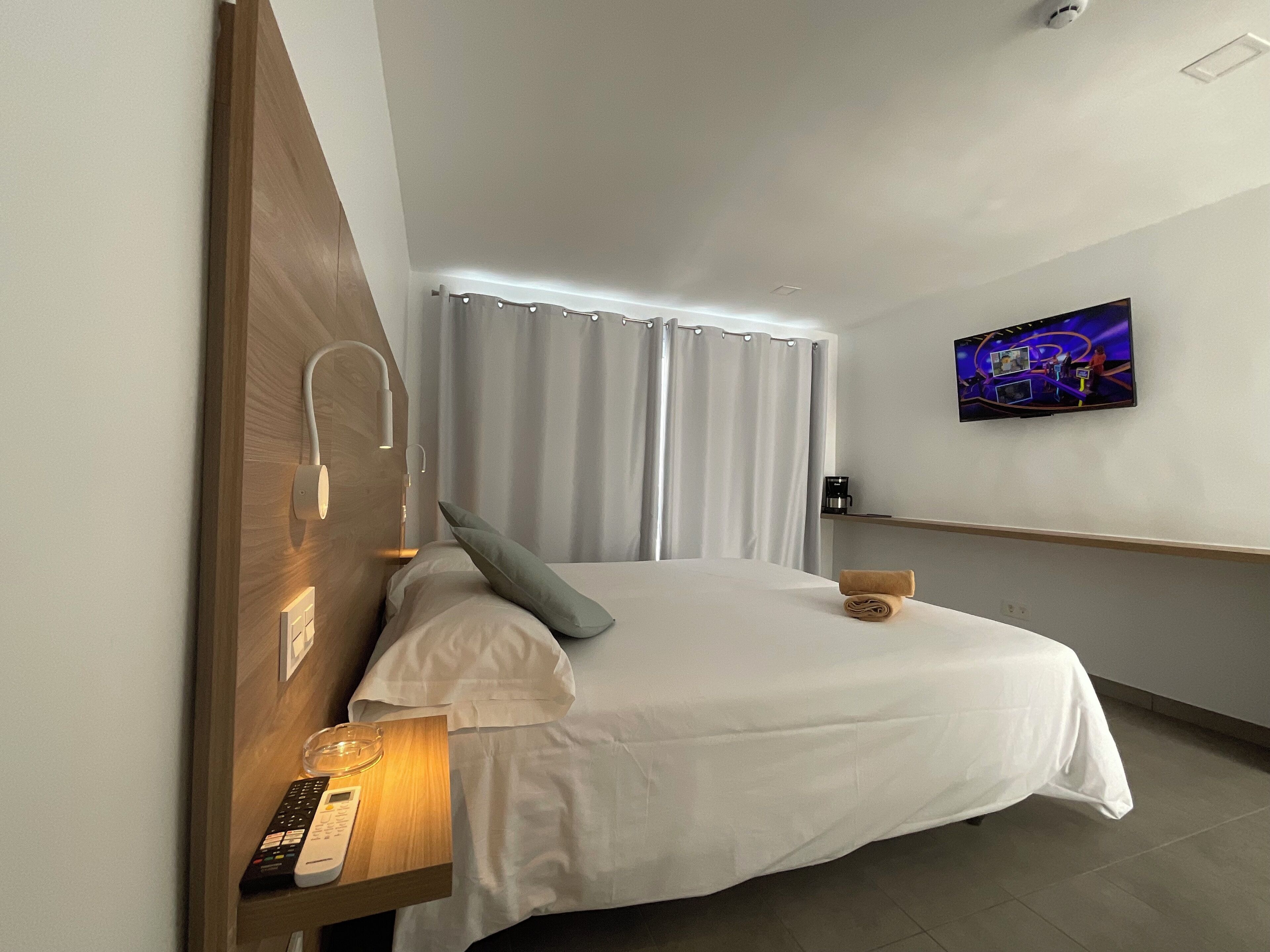 A hotel room with a bed, a TV mounted on the wall, a bedside table with a lamp, and curtains.