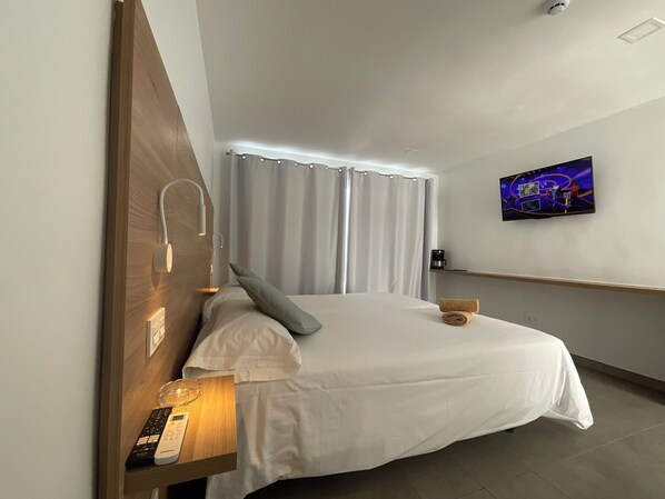 Une chambre d’hôtel avec un lit, un téléviseur fixé au mur, une table de chevet avec une lampe et des rideaux.