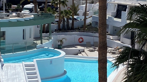 A swimming pool with lounge chairs and umbrellas, surrounded by palm trees and modern buildings. - Kikerestudios (Puerto del Carmen)