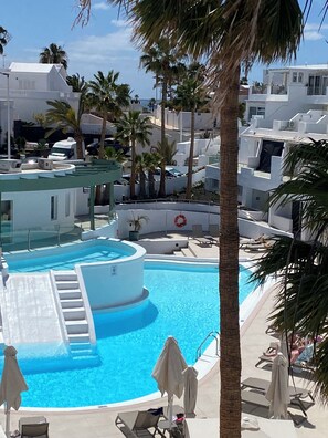 A swimming pool with lounge chairs and umbrellas, surrounded by palm trees and modern buildings. - Kikerestudios (Puerto del Carmen)