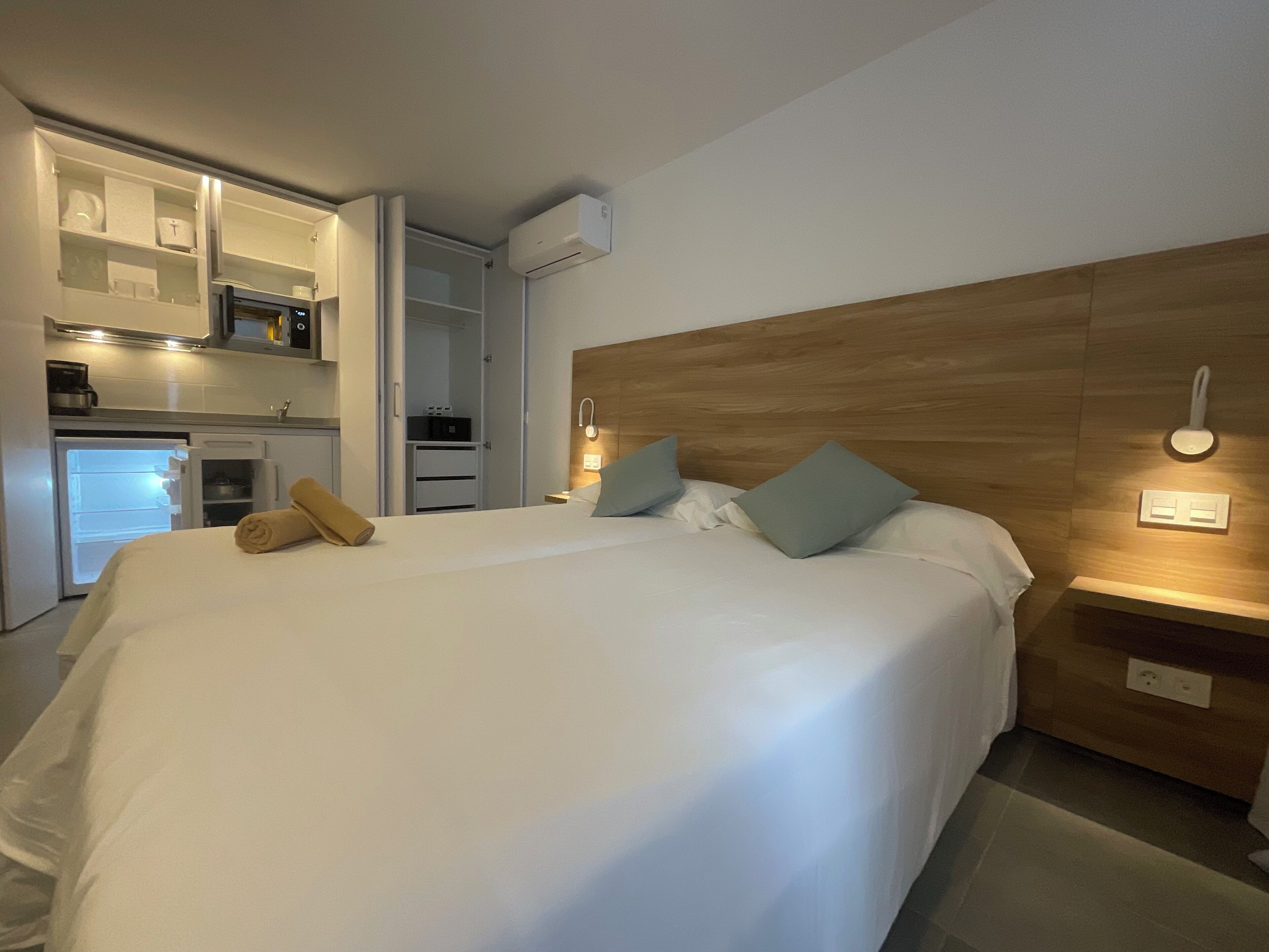 A hotel room with a bed, a wooden headboard, a nightstand, a bedside table, a wardrobe, a built-in kitchen with a refrigerator, and an air conditioning unit.