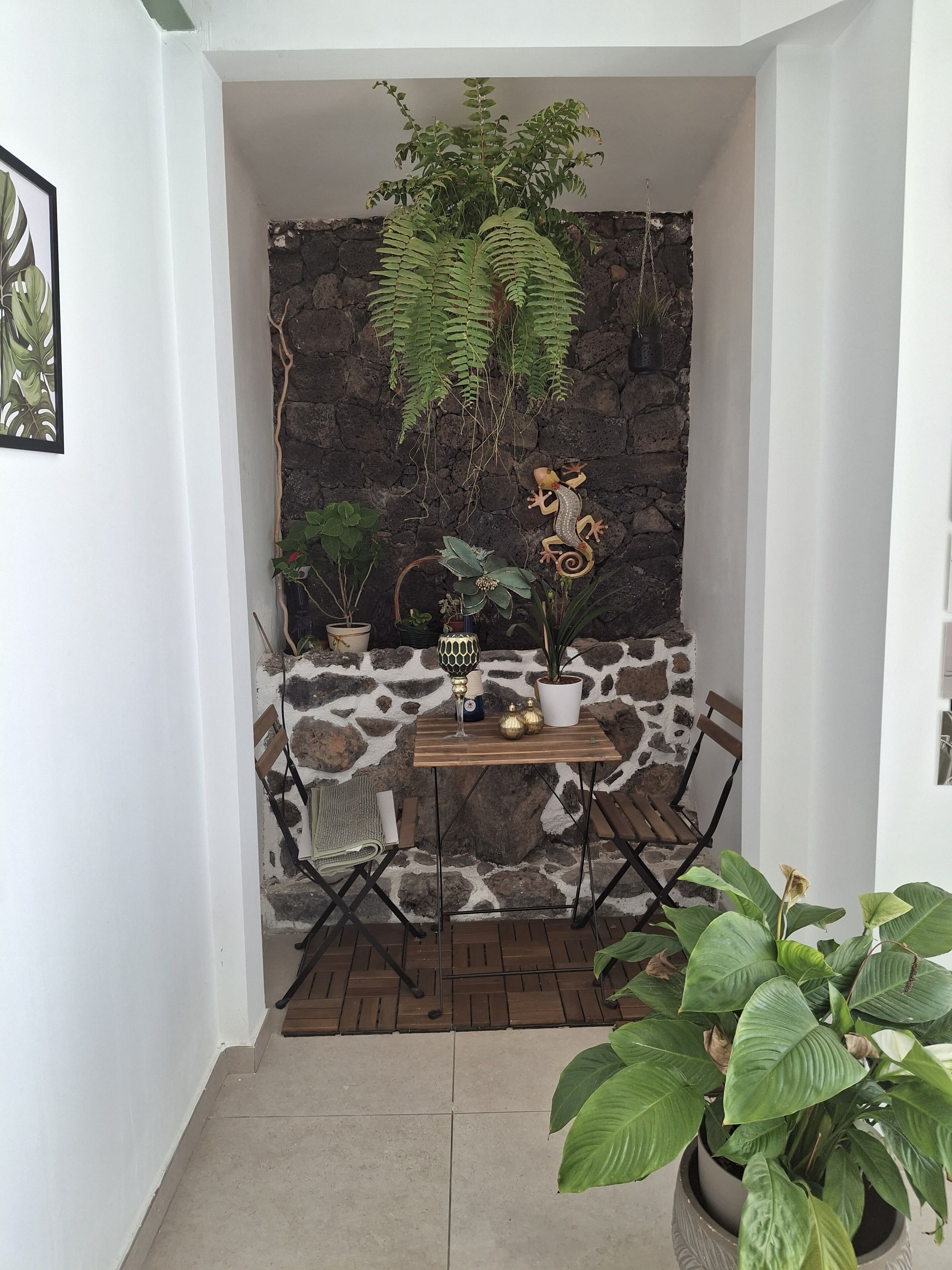 Un rincón pequeño y acogedor con un fondo de pared de piedra, una mesa de madera con una lámpara decorativa y varias plantas en macetas.