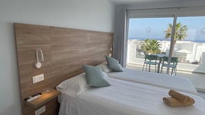 A hotel room with two beds, a wooden headboard, a small table with chairs, and a view of the sea and buildings. - Kikerestudios (Puerto del Carmen)