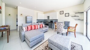 A modern living room with a grey sofa, a dining area with a table and chairs, a kitchen with white cabinets, and a large window providing natural light. - Ada 5 (Bedfordview)