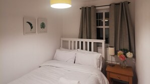 Un dormitorio con una cama individual, una mesita de noche con lámpara, una ventana con cortinas y dos cuadros en la pared.