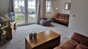 Una sala de estar con un sofá marrón, una mesa de centro de madera con dos tazas, un sillón informal gris y un televisor sobre un mueble de madera.