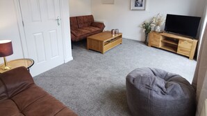 Una sala de estar con un sofá marrón, un sillón de peluche gris, una mesa de centro de madera, un mueble para televisor y una puerta blanca.