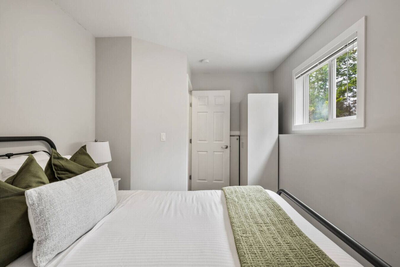 Une chambre à coucher avec un lit, des oreillers, une table de chevet, une lampe et une fenêtre donnant sur des arbres.