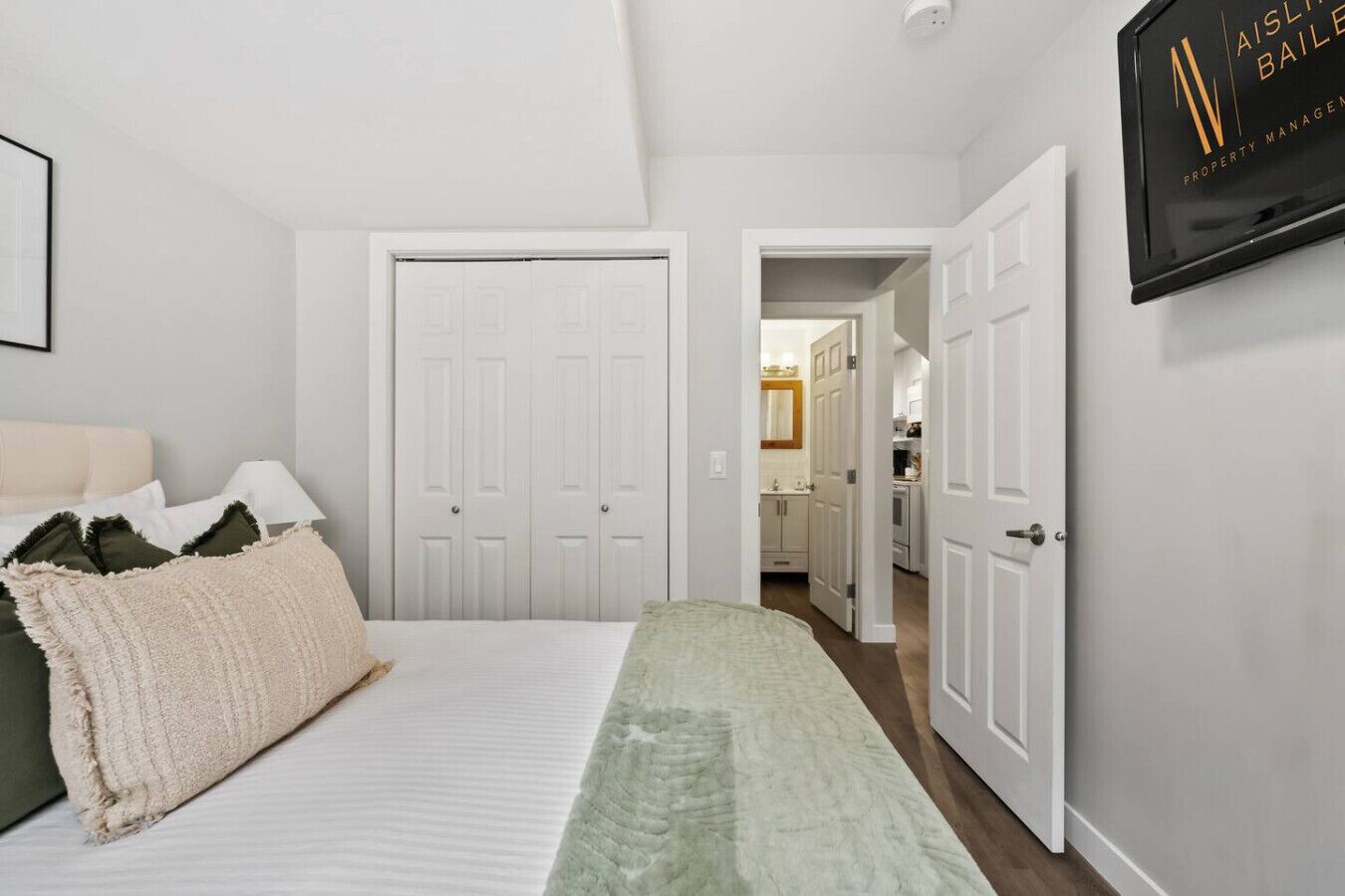 A bedroom with a bed, pillows, a bedside table, and a wardrobe. There is a hallway leading to a bathroom and a kitchen.