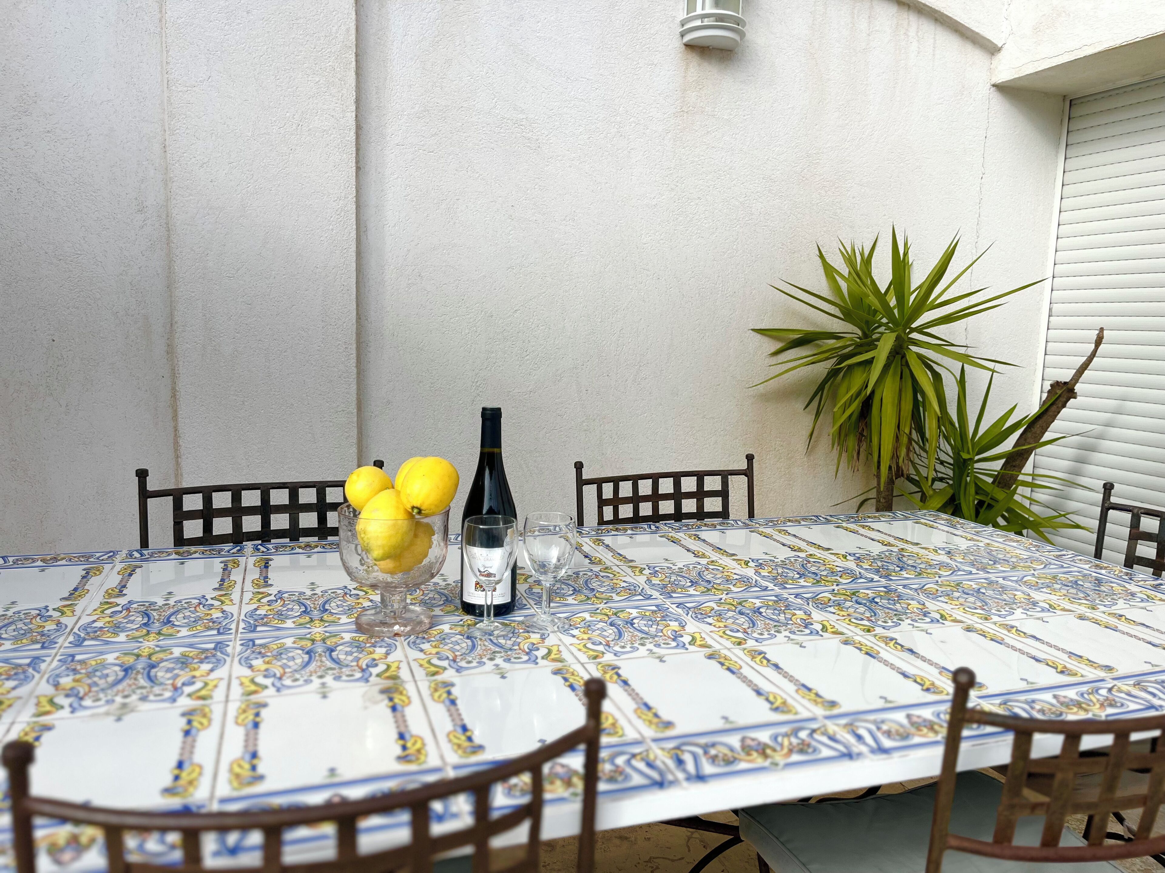 Une table ronde recouverte d’une nappe à motifs, entourée de chaises en bois. Un bol rempli de citrons et une bouteille de vin sont posés sur la table.