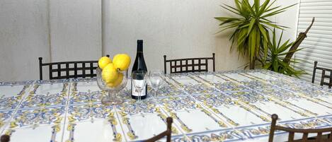 A round table with a patterned tablecloth, surrounded by wooden chairs. A bowl with lemons and a bottle of wine are placed on the table.