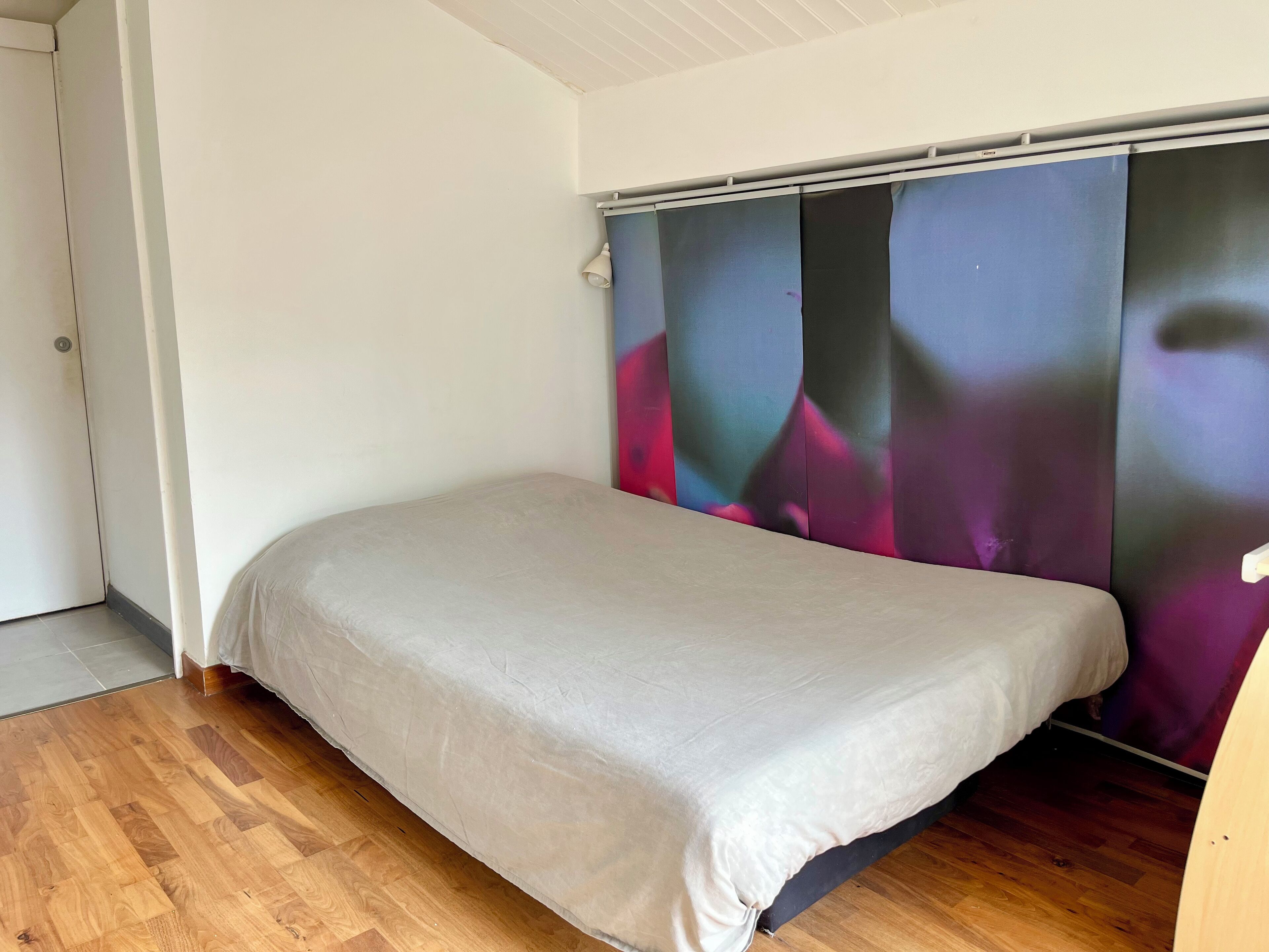 A small, single-bed room with a wooden floor, a plain white wall, and a large window with a view of colorful buildings.