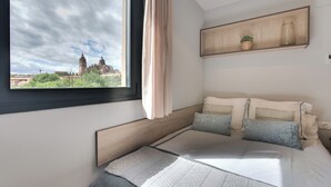 A bedroom with a large window, a bed with patterned bedding, a wooden headboard, a shelf with a plant and a jar, and a view of a historic building. - Livensa Living Salamanca Parking Free (Salamanca)