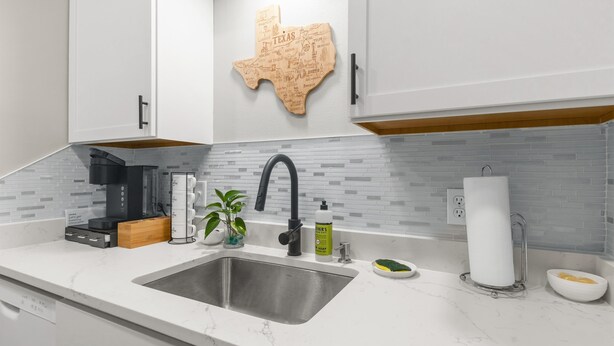 A modern kitchen with a white countertop, a stainless steel sink, a coffee maker, a plant, and a Texas-themed wall decoration. - Camellia: Studio Apt, 4 Minute walk to A&M (College Station)