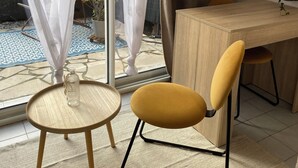 Una habitación con una mesa de madera para comer, una silla amarilla, una mesita redonda con una botella y cortinas translúcidas con motivos de hojas.