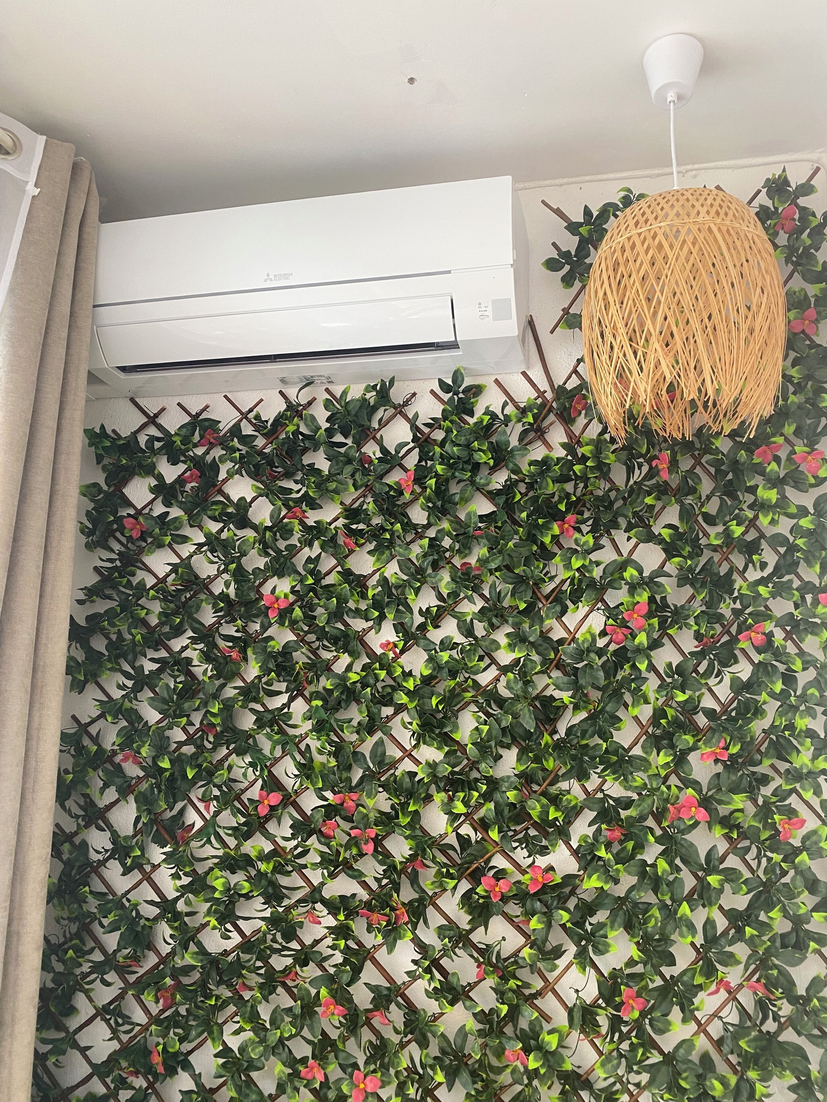 A room with a white air conditioning unit, a woven pendant light, and a wall covered with a green leafy climbing plant.