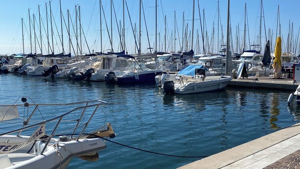 Un puerto deportivo con varias embarcaciones amarradas, un paseo marítimo pavimentado y un cielo azul despejado.