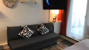 Una habitación de hotel moderna con un sofá oscuro, dos cojines estampados, un reloj de pared, un televisor de pantalla plana, una mesita con un objeto decorativo y una ventana con cortinas.