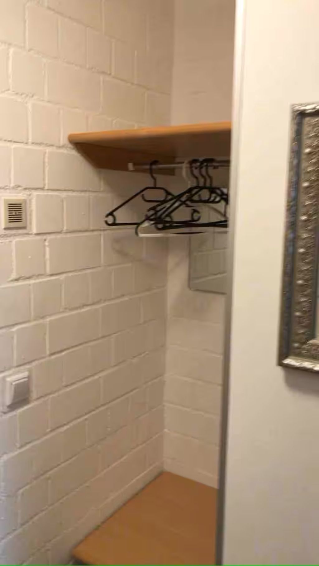 A corner of a room with a white brick wall, a wooden shelf, and a metal hanger rack.