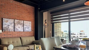 A modern living room with a brick wall, a sofa, a patterned armchair, a small table with a vase, and a view of buildings through large windows. - 1 Bed With Ocean Front Views in the Romantic Zone - C205 (Puerto Vallarta)