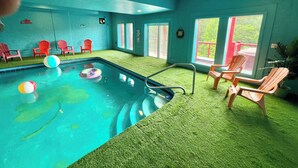 An indoor swimming pool with artificial grass, surrounded by red chairs and a wooden deck.