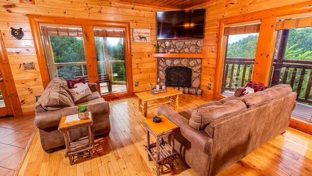 A log cabin interior with a stone fireplace, two sofas, a coffee table, and a TV mounted above the fireplace.