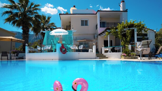 A swimming pool with inflatable flamingos, a white building with balconies, and outdoor seating. - Aegli Hotel Studios (Thasos)