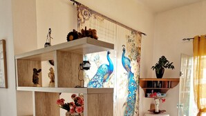 A living room with a wooden bookshelf, a sofa with a yellow cover, a peacock mural, and a patterned rug. - Aegli Hotel Studios (Thasos)