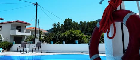 A swimming pool with a lifebuoy and umbrella, surrounded by a tiled deck and a building with a red roof.