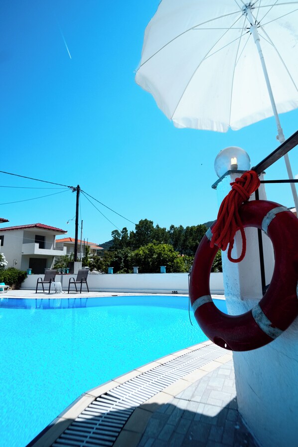 A swimming pool with a lifebuoy and umbrella, surrounded by a tiled deck and a building with a red roof.