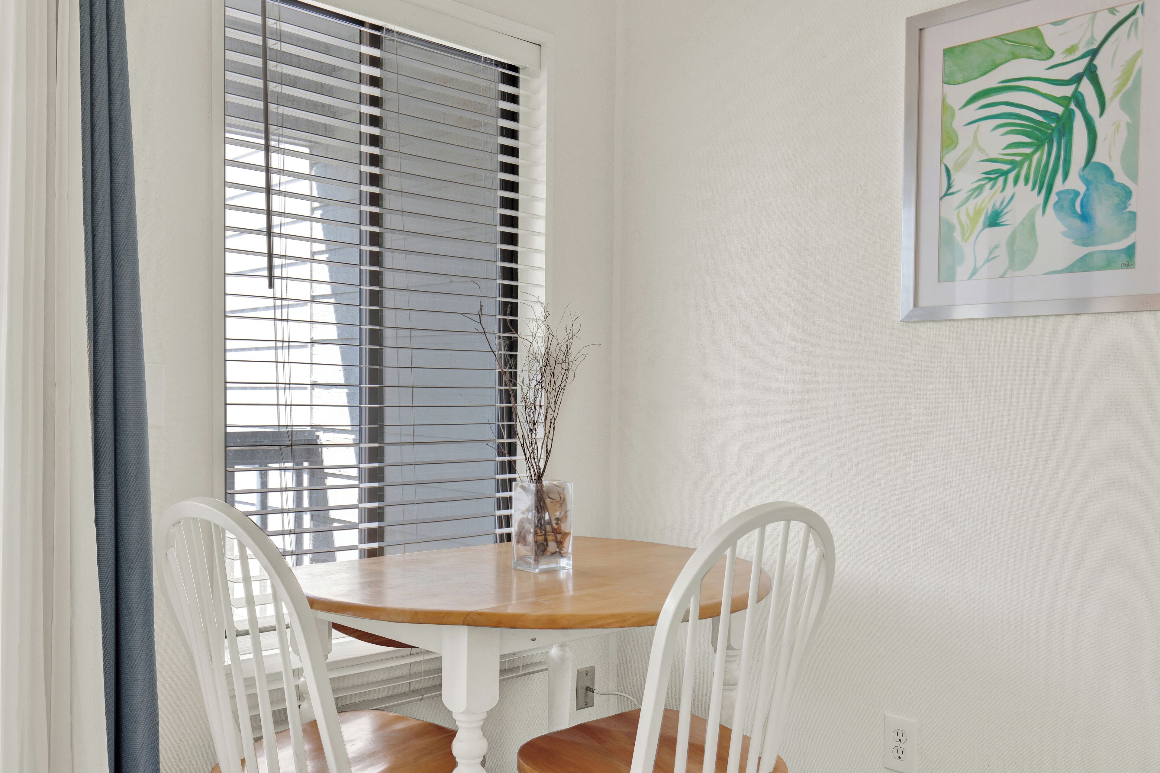 A dining area with a wooden table and white chairs, a vase with branches, and a framed botanical print on the wall.