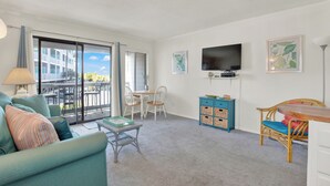 A living room with a sofa, a table, a TV, and a balcony with a view of buildings. - Charming 1-bedroom condo in serene Tybee Island (Tybee Island)