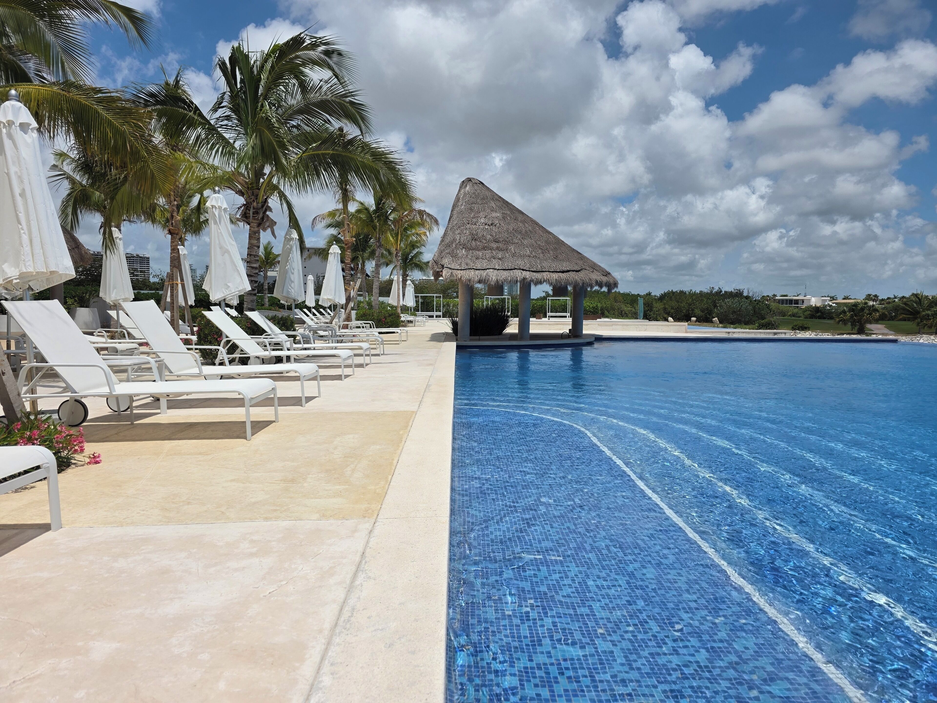 Un espace aménagé au bord de la piscine, avec des chaises longues et des parasols, une structure à toit de chaume et une piscine à l’eau d’un bleu limpide.
