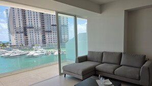 A modern living room with a grey sectional sofa, a dark wood coffee table, and a glass door leading to a balcony with a view of a marina and high-rise buildings. - Marea Puerto Cancun, luxury+comfort+the best view (Cancún)