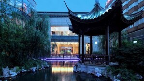 A traditional pavilion with a roof, a water feature, and a building with glass windows in the background.