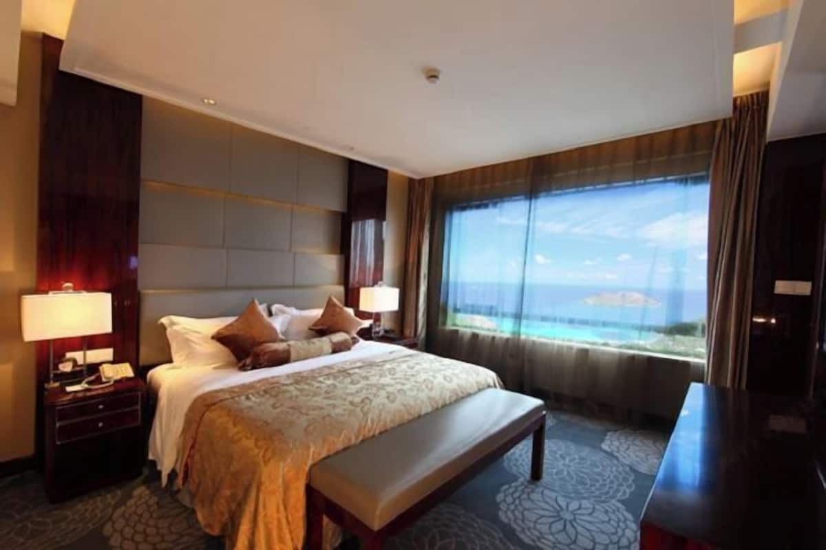Habitación de hotel con una cama grande, un banco, una mesita de noche y vista al océano.