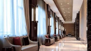 A hotel lobby with a row of sofas and armchairs, a long corridor, and a large window with curtains.