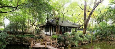 A traditional pavilion over a pond with a wooden bridge, surrounded by lush greenery.
