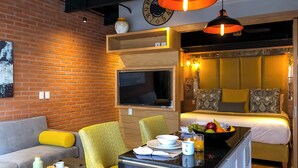 A modern hotel room with a brick wall, a flat-screen TV, a bed with a yellow headboard, a dining area with a table and chairs, and a small kitchen area.
