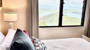 Habitación de hotel con un gran ventanal que ofrece vistas a un lago y colinas. Hay una cama con almohadas blancas y rojas, y una lámpara en la mesita de noche.