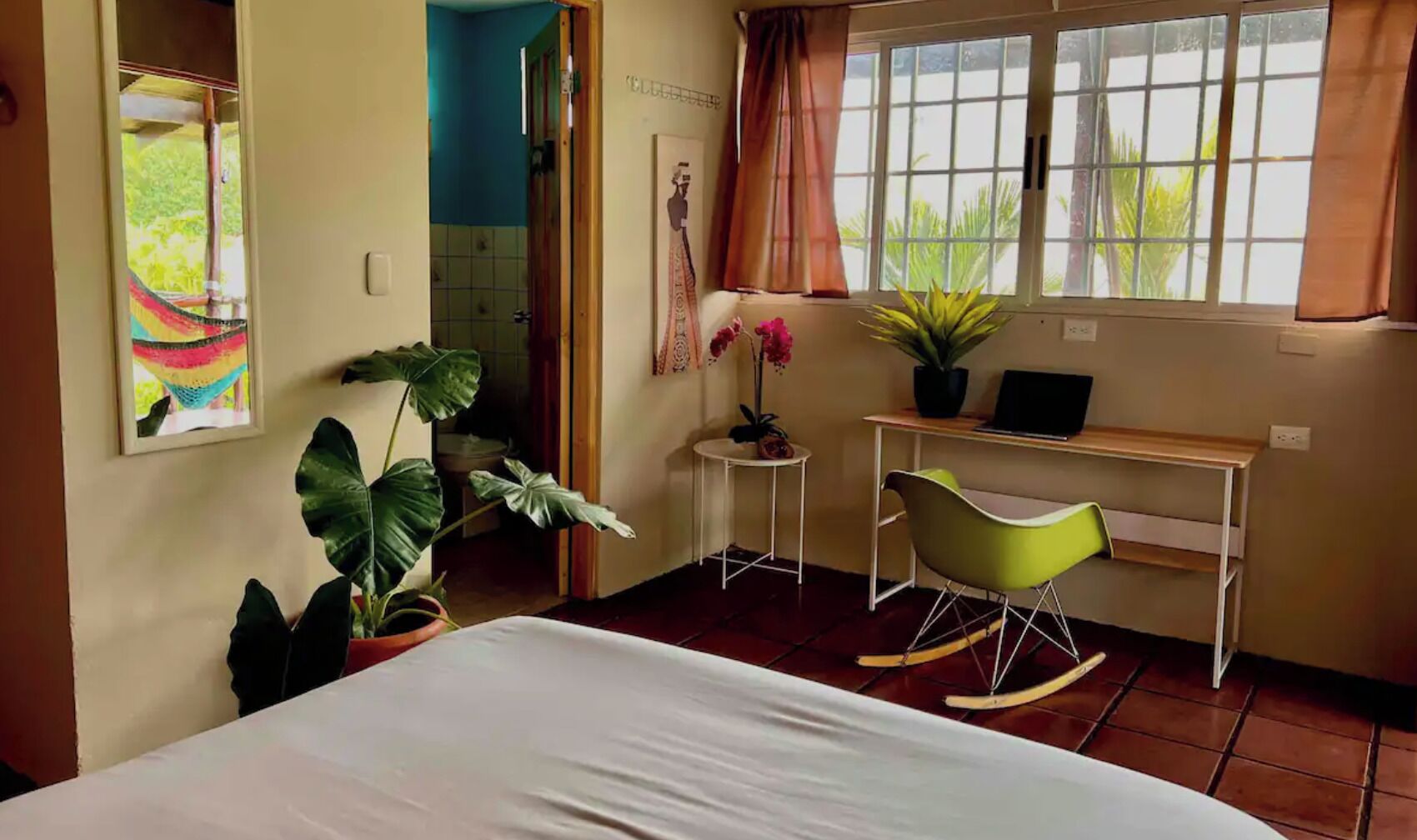 Une chambre à coucher avec un lit, un bureau, un fauteuil à bascule, une plante en pot et une fenêtre avec des rideaux.