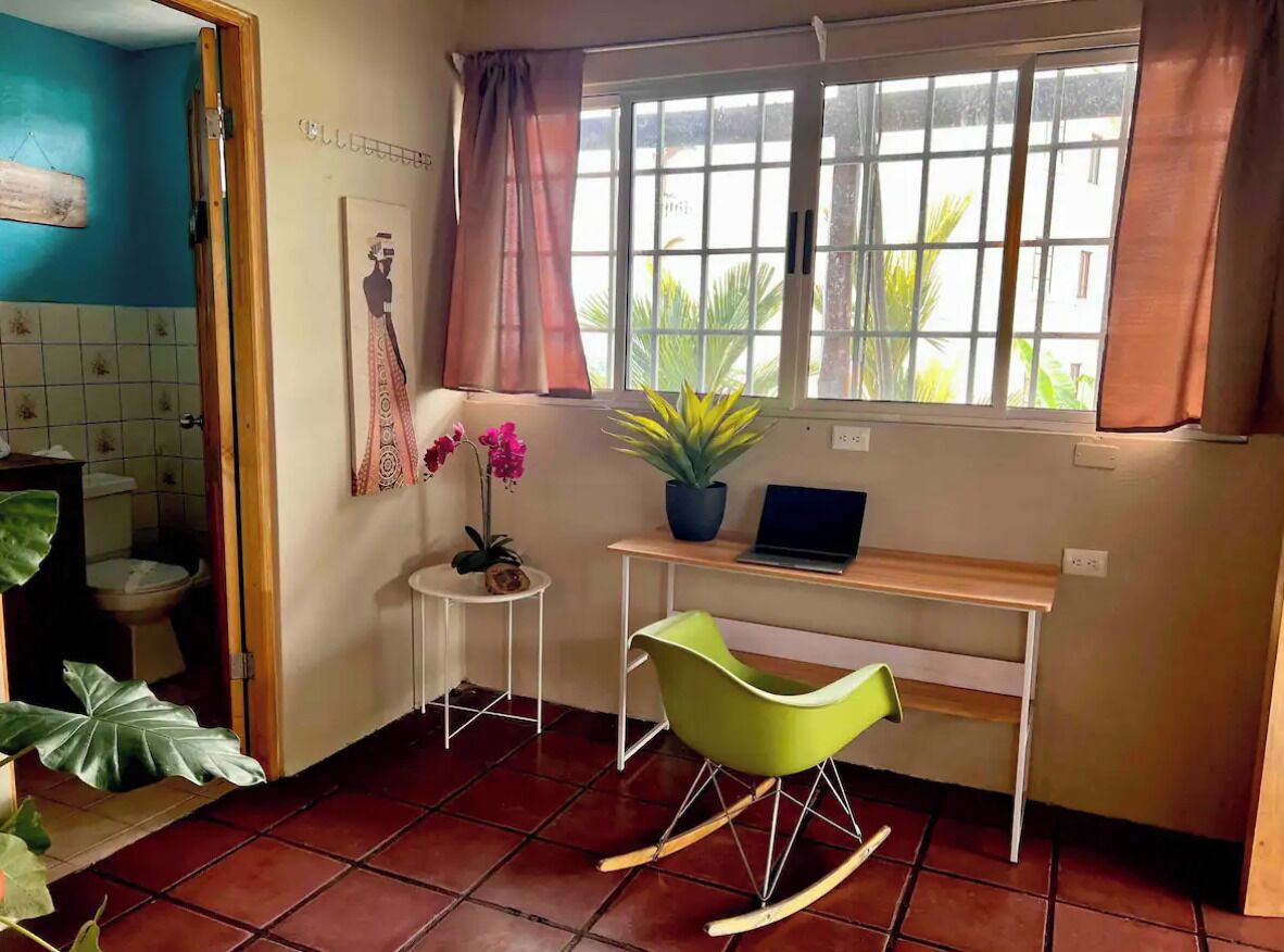 Ein kleines Zimmer mit Schreibtisch, Schaukelstuhl, Blumentopf und einem Fenster mit Vorhängen.