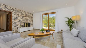 A modern living room with a stone wall, a large window, a sofa, a coffee table, and a TV. - Villa Es Garrover (Alcúdia)