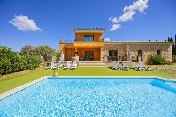 A two-story house with a swimming pool, a garden with lounge chairs, and a stone facade.