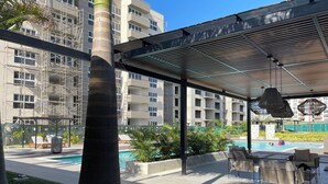 Zona de piscina en la azotea con sillones de descanso, un área de estar cubierta y vista a un edificio en construcción.