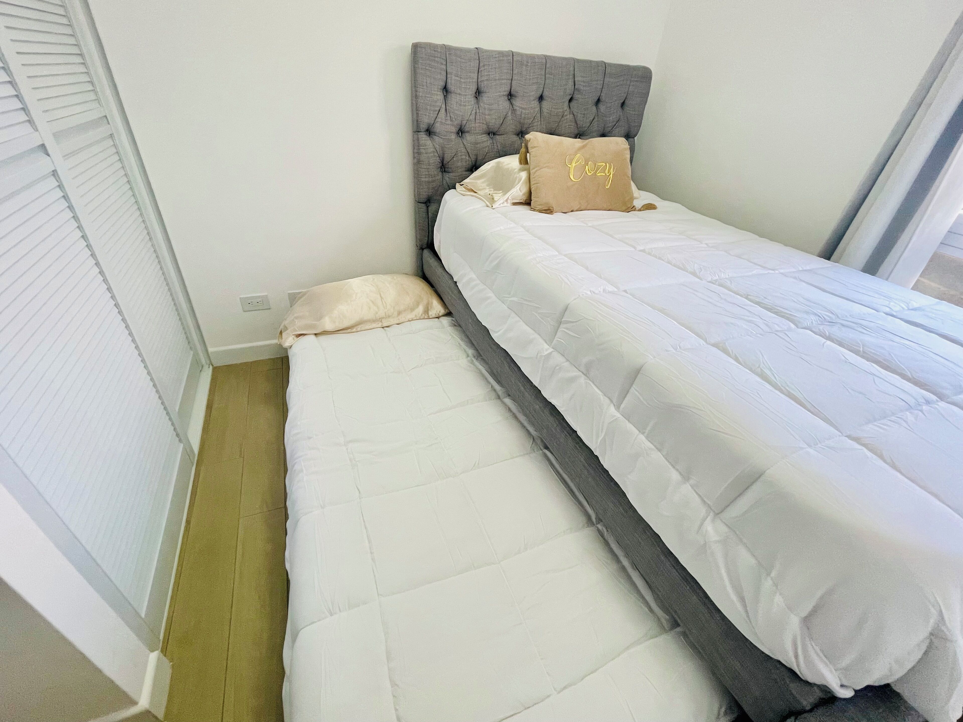 Et lille soveværelse med en grå polstret sengegavl, en hvid madras og en beige pude.