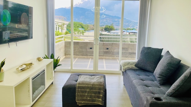 Una sala moderna con un sofá gris, un mueble de TV blanco y una televisión grande.