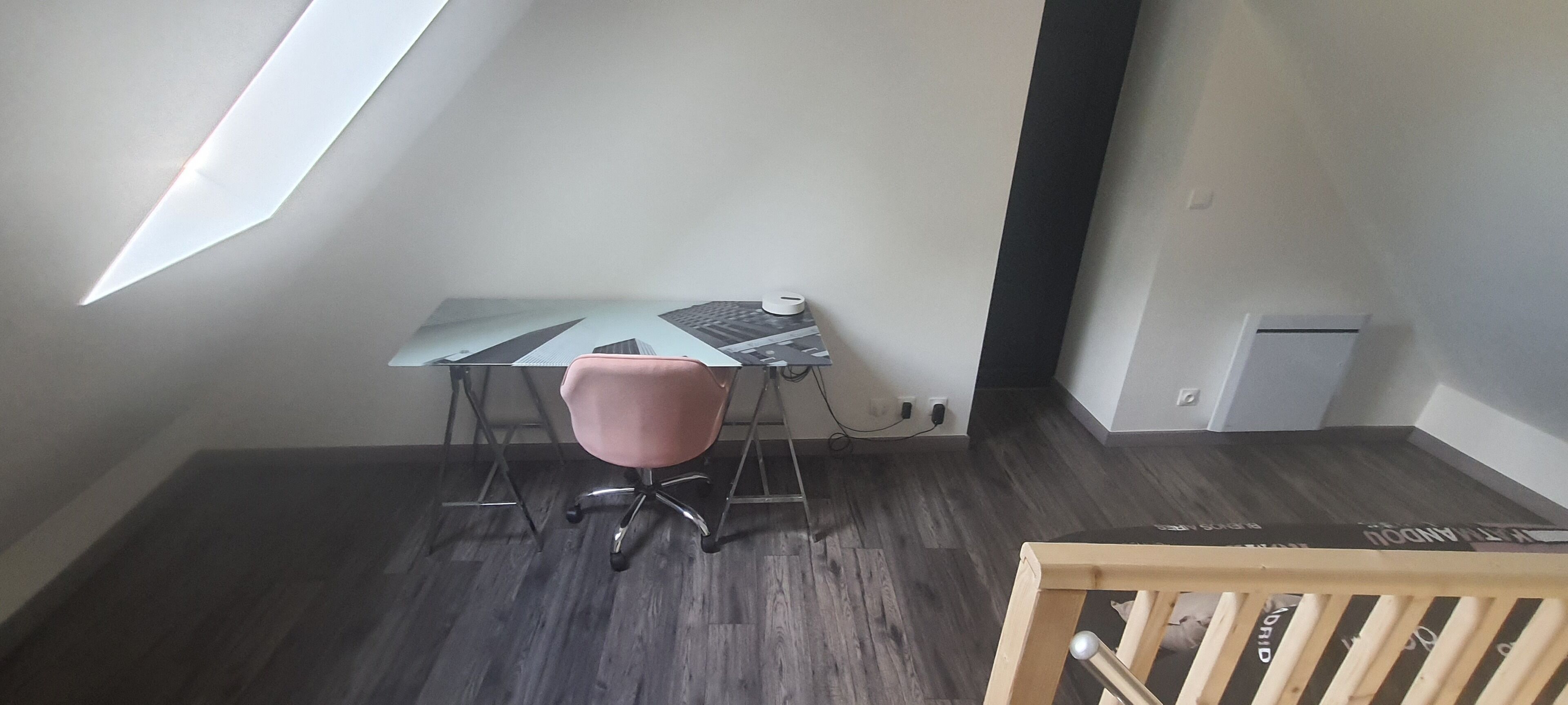 A small, modern room with a slanted ceiling, a desk with a computer, a pink chair, and a crib.